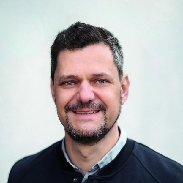 Mann mit dunklem Haar trägt ein schwarzes Hemd über einem hellen Hemd vor hellem Hintergrund