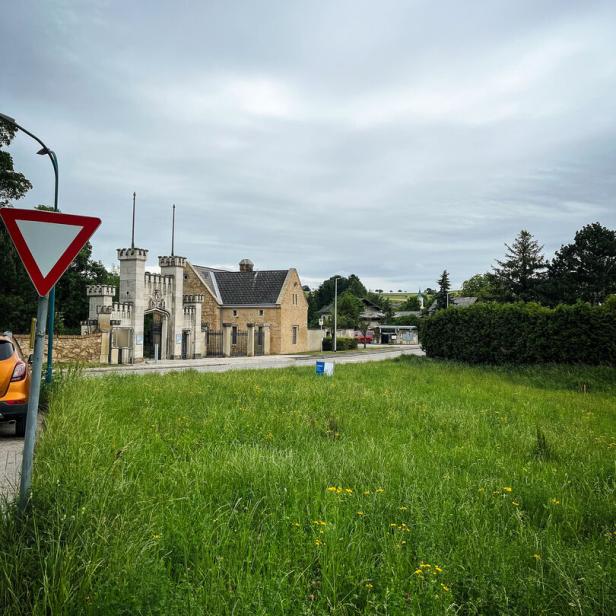 Ein altes Gebäude mit Türmen steht hinter einer Wiese, links ist ein Vorfahrt-gewähren-Schild und ein orangefarbenes Auto zu sehen.