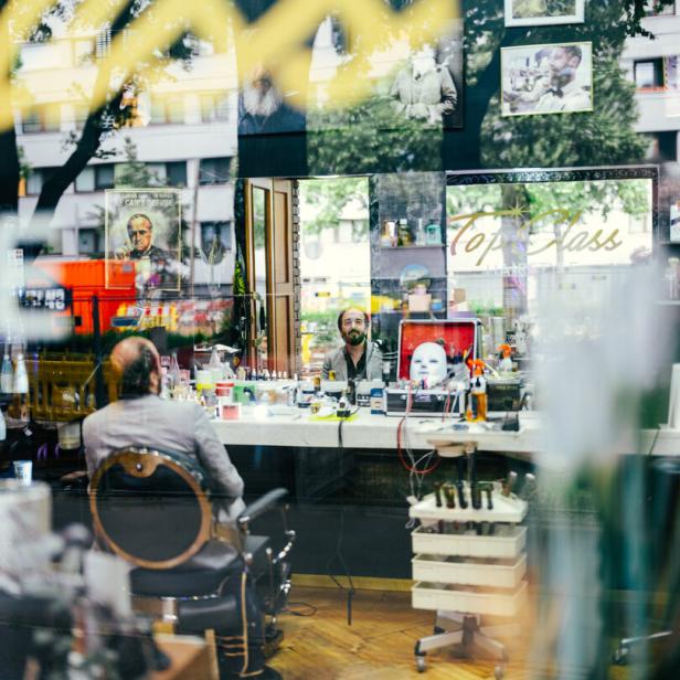 Ein Mann sitzt vor einem Spiegel im Friseursalon „Top Class Hairstyle“, während sich sein Gesicht in der Scheibe spiegelt.