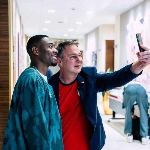 David Alaba lächelt, während ein Mann in einem roten Trikot und blauem Sakko, Sportminister Babler, ein Selfie mit ihm in einem Flur macht.