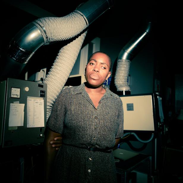 Die Künstlerin Onyeka Igwe blickt ernst, mit leicht geneigtem Kopf, aus einem halbdunklen Raum in die Kamera, im Hintergrund große Röhren, wie in einem Maschinenraum
