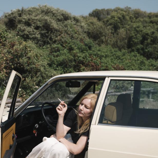Die Schauspielerin Sandra Hüller mit blonder Perücke, rauchend, in einem geparkten Auto sitzend, im Hintergrund ein Wald