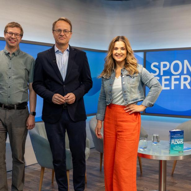 Drei Personen stehen vor einem blauen Hintergrund mit der Aufschrift 'SPONTAN GEFRAGT', links und rechts je ein grauer Sessel und ein runder Glastisch mit zwei Gläsern und einem Buch