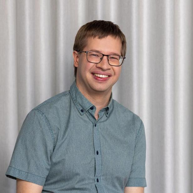 Mann mit kurz geschnittenem Haar und Brille sitzt vor einem hellen Vorhang und trägt ein kurzärmeliges Hemd