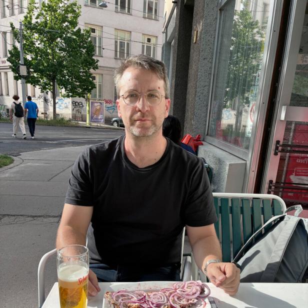 Ein Mann mit Brille und schwarzem T-Shirt sitzt vor einem Schmalzbrot und einem großen Bier