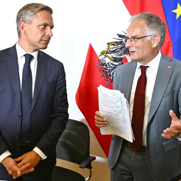 Zwei Männer im Anzug, die Minister Hattmannsdorfer und Marterbauer, diskutieren vor österreichischer und EU-Flagge, einer hält Unterlagen in der Hand.