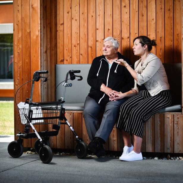 Eine junge Frau spricht tröstend mit einer älteren Frau, die mit Rollator auf einer Holzbank sitzt.